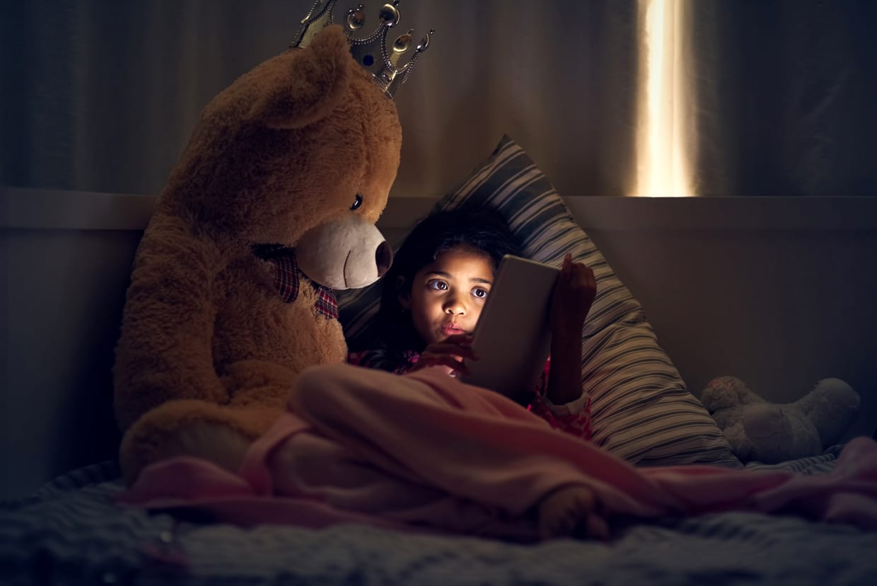 A little girl using a tablet while lying in bed with her teddy bear at night.